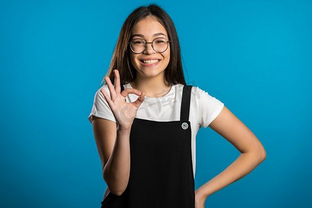 年轻的亚洲女性，有着完美的妆容，在蓝色背景上做着“OK”的手势。