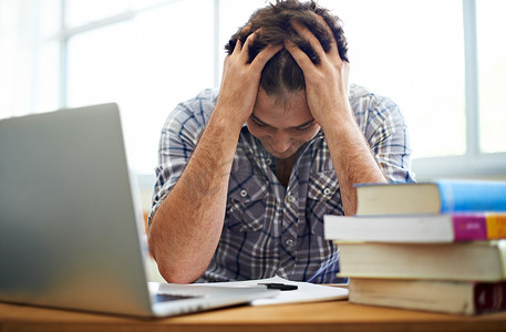 考试压力越来越大……一名学生双手抱头坐在一堆课本和笔记本电脑旁边。