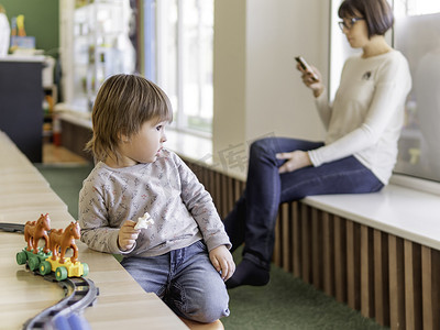 蹒跚学步的孩子在玩五颜六色的玩具积木，而他的母亲或保姆则在智能手机上发短信。