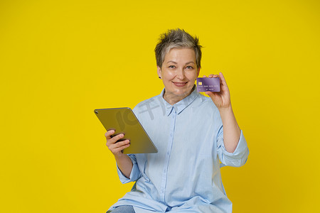 头发灰白的成熟女性手里拿着信用卡、借记卡和平板电脑，进行在线支付或在线购物，隔离在黄色背景中。