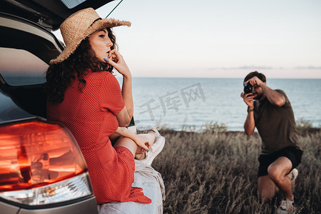 水里的衣服摄影照片_男人拍摄坐在海背景下打开的汽车后备箱里的穿着红色衣服和帽子的女人
