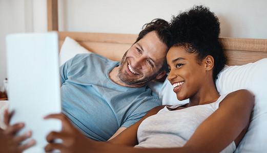 阅读app界面摄影照片_快乐、放松和无忧无虑的异族情侣在数字平板电脑上微笑着躺在床上自拍。