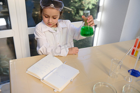 绿色阅读摄影照片_迷人的小女孩化学家一边阅读化学教科书中的化学公式，一边摇动装有绿色溶液的烧瓶