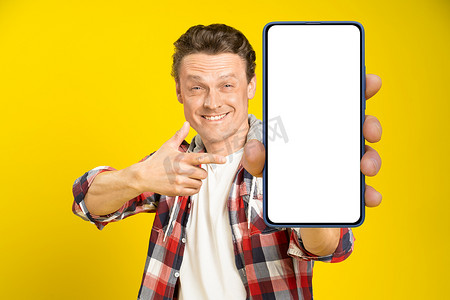 迷人的快乐男人指着带有白色空屏幕的智能手机，身穿红色格子衬衫和牛仔裤，手机显示屏模拟在黄色背景中隔离。