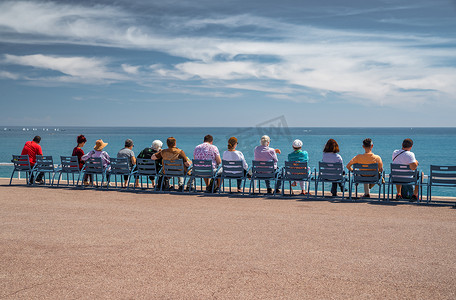 人们坐在法国尼斯堤防上著名的蓝色椅子上，看着利古里亚海的蔚蓝海水、宁静、晚年、休息、老年人、地平线、社会