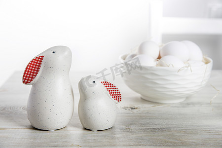 在一个白色的斯堪的纳维亚风格的厨房里，两个白色野兔的小雕像，在木锯上有大大小小的红耳朵，还有一个背景是鸡蛋的碗