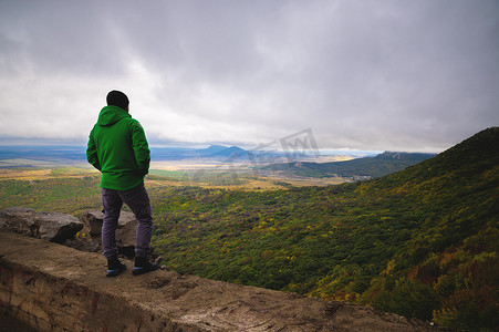 一个徒步旅行的人，穿着一件鲜艳的夹克站在一座山顶上，眺望远方的山脉和多云的天空