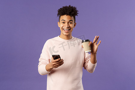 快乐的年轻男学生拿着咖啡和手机的肖像，挥手打招呼，微笑友好地打招呼，在大学校园附近与同学见面，紫色背景