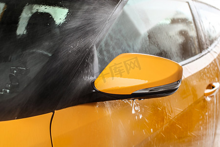 黄色汽车的侧镜在自助洗车中清洗，高压喷水，水滴飞来飞去。