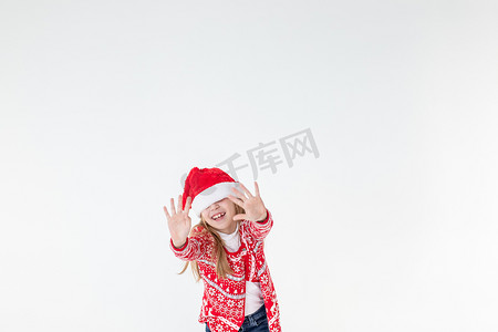快乐的女孩跳舞的画像，穿着红色圣诞毛衣和孤立在白色背景上的圣诞老人帽子。穿着圣诞马鹿毛衣的年轻白种人快乐微笑的金发女孩很开心。