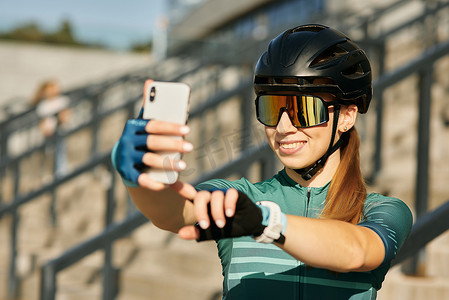女自行车运动员摄影照片_穿着自行车服装和防护装备的快乐职业女自行车手在使用智能手机自拍时微笑，在市中心骑自行车后休息