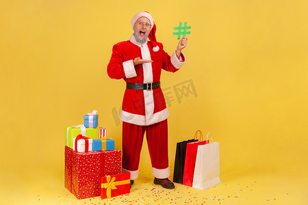 社区老人摄影照片_留着胡子的老人的全长肖像，穿着圣诞老人服装，周围环绕着圣诞礼物，指着手中的绿色标签。