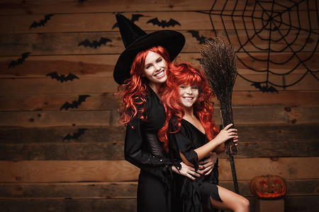 巫婆背景摄影照片_美丽的白人母亲和她的女儿，长着红色的长发，穿着巫婆服装庆祝万圣节，在木制工作室背景上摆出蝙蝠和蜘蛛网的姿势。
