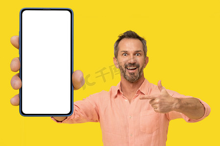 中年英俊的灰发男子指着手中巨大的智能手机，惊讶地微笑着看着穿着黄色隔离桃色衬衫的相机。