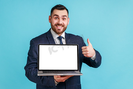 留着胡子摄影照片_乐观高兴的留着胡子的男人穿着官方风格的西装，拿着笔记本电脑，竖起大拇指，表示认可，对互联网服务感到满意。