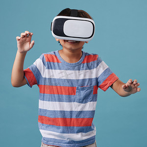 男孩站着摄影照片_年轻的混血男孩站着，戴着无线 vr 耳机，在蓝色背景下玩电子游戏。