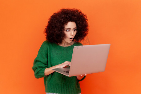 身穿绿色休闲风格毛衣、留着非洲发型、在便携式电脑上工作、张开嘴看着显示屏的震惊女性的肖像。