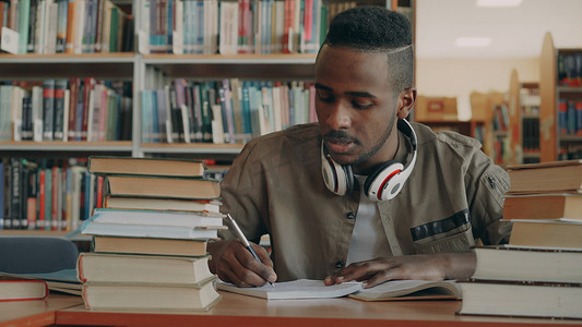 美国帅哥摄影照片_积极的非洲裔美国年轻帅哥，戴着大耳机，坐在桌边，拿着书，在抄写本上写字
