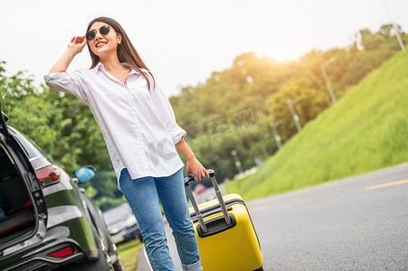 拖着行李旅行摄影照片_戴着墨镜的亚洲美女在公路旅行中独自拖着黄色手提箱行李。