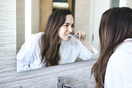 刷牙摄影照片_迷人的白人白人女性穿着白色长袍在浴室镜子前刷牙的生活方式照片