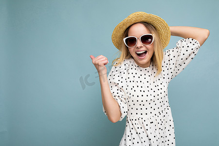 可爱蓝色摄影照片_年轻漂亮可爱快乐的金发女人穿着休闲服、太阳镜和草帽的照片在蓝色背景中被隔离，有复制空间并庆祝胜利。