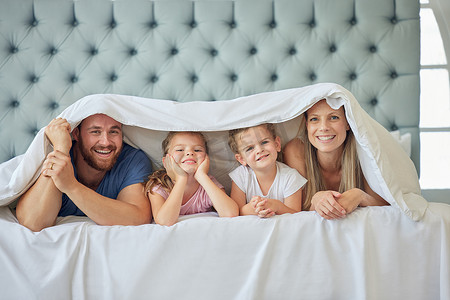 快乐的父母和小孩躺在家里的床上，盖着毯子的肖像。