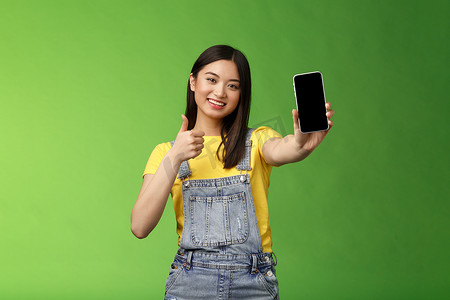 开朗可爱的亚洲女性判断优秀的智能手机游戏，展示大拇指标志，伸出手臂握住电话屏幕，展示出色的应用程序，推广应用程序，站立绿色背景