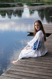 一个穿着乌克兰刺绣衬衫的女孩坐在码头上，湖水中倒映着云彩。
