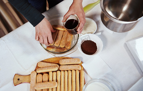 马斯卡彭摄影照片_女手在制作自制提拉米苏蛋糕时，将手指饼干浸在盘子上的甜浓咖啡中，顶视图