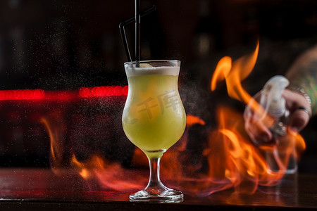 点燃火焰摄影照片_酒保在吧台上洒上带亮绿色冷鸡尾酒的照明玻璃，并在上面点燃火焰