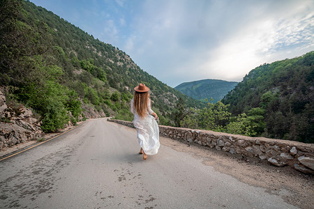流浪着摄影照片_穿着棕色帽子和白色连衣裙的女旅行者看着令人惊叹的山脉和森林、流浪旅游概念、大气的史诗时刻