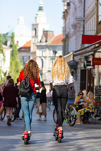 公共生活摄影照片_在城市环境中乘坐公共租赁电动滑板车的时尚时尚少女的后视图。