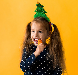 肖像正面开朗微笑快乐的小女生女孩圣诞树装饰波尔卡圆点连衣裙咬吃橙色背景上的橙色胡萝卜。
