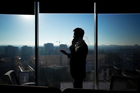 窗户背景的现代办公室里一个商人的剪影，一个男人在电话里说话