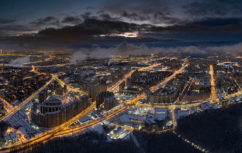 圣彼得堡-俄罗斯黄昏时的空中冬季城市景观，无人机飞过巨大的住宅区和公园，建筑起重机，夜间照明，云彩漂浮在无人机下方