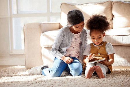两个混血小姐妹坐在地板上，一起在家里玩数字平板电脑。