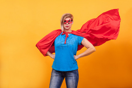 身穿超级英雄服装的年轻女性在黄色背景中骄傲地站在空中，身着红色斗篷
