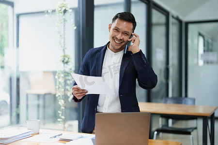 一位亚洲男性企业主微笑着与合作伙伴通电话，谈判合资企业以与另一家公司竞争的肖像