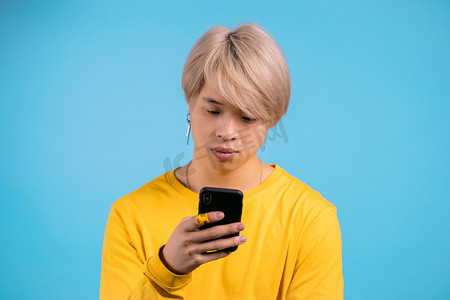 英俊的韩国男子在蓝色工作室墙上使用智能手机。
