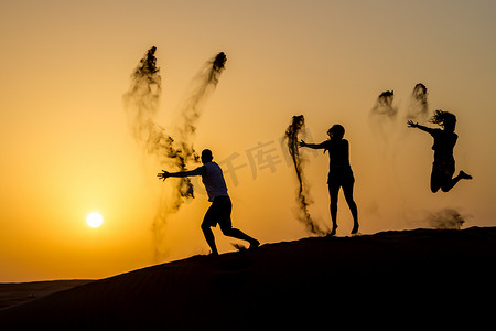 金色日落时分，快乐旅行的人在沙丘上跳跃，向空中抛沙的剪影