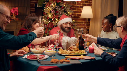 快乐的男人伪装成圣诞老人，与家人一起享用圣诞晚餐，碰杯香槟