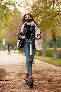 在科维德大流行期间，戴着防护面罩的休闲白人青少年在城市公园骑城市电动滑板车。