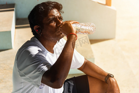特写亚洲年轻运动跑步者黑人戴着运动员耳机，他在户外街头健康公园跑步后从瓶子里喝水，健康的运动锻炼概念