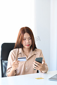 年轻的亚洲女性手持信用卡并使用智能手机进行网上购物。