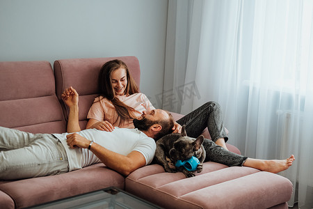 成年夫妇在家里和法国斗牛犬一起休闲，快乐的女人和男人在粉红色的沙发上和宠物一起放松