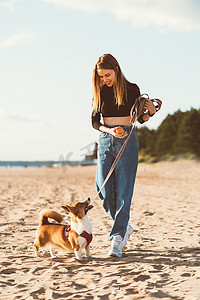 美丽的年轻女子和 Corgi 小狗一起玩耍，沿着海洋海滩散步。