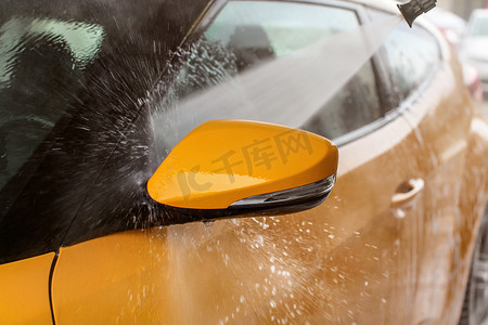 黄色汽车的侧镜在自助洗车中清洗，喷射的水击中车窗，水滴飞向四面八方。