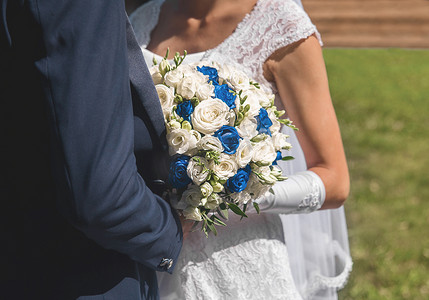 身着白色礼服的新娘和新郎站在彼此旁边拥抱，手捧玫瑰花婚礼花束在公园散步