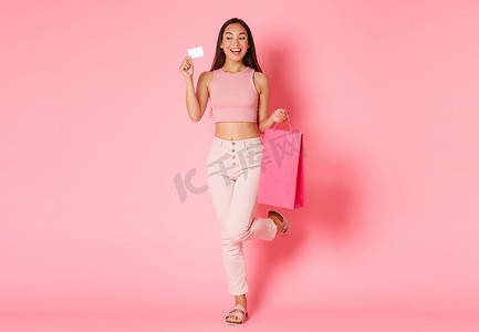所有消费者摄影照片_全长华丽的梦幻般微笑的亚洲女孩把所有的钱都浪费在银行账户上买新衣服，拿着购物袋和信用卡，在粉红色的背景下看起来很乐观