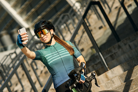 穿着自行车服装和防护装备的快乐职业女自行车手在使用智能手机自拍时微笑，在市中心骑自行车后站立和休息